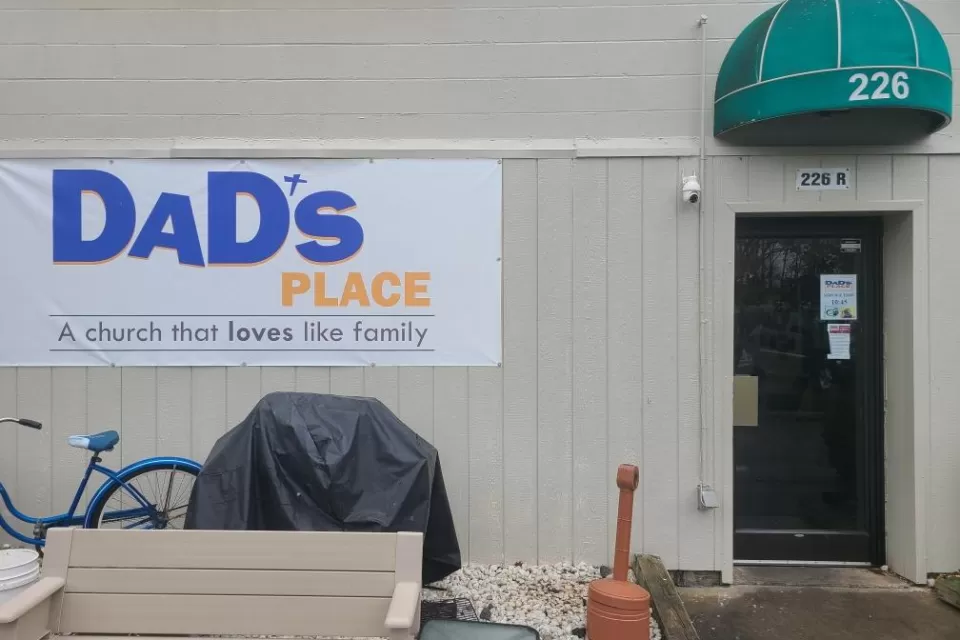 White sign on side of tan building with metal siding and green overhang, reading "Dad's Place, a church that loves like family."